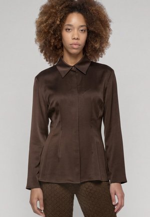 Femme aux cheveux bouclés portant un chemisier en satin marron foncé à manches longues boutonné et un pantalon marron texturé sur un fond uni.