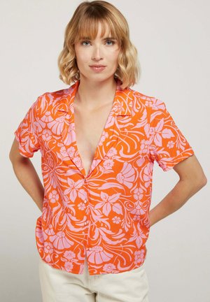 Femme aux cheveux blonds courts portant une chemise orange à manches courtes avec des motifs floraux roses et un pantalon blanc, debout les mains sur les hanches.