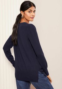Granatowy sweter z długim rękawem o żebrowanej strukturze, z bocznymi szczelinami i luźnym krojem, zestawiony z ciemnymi dżinsami. Prosty fason z okrągłym dekoltem.