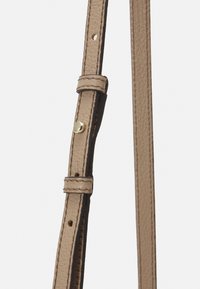 Beige nahkahihna, jossa on teksturoitu pinta, säädettävä solki ja kaksi rivistöä ompeleet. Kultaiset yksityiskohdat hihnassa.