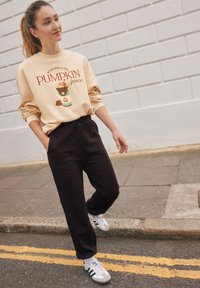 Sweatshirt beige avec le graphisme "PUMPKIN spice", associé à un pantalon de survêtement noir et des baskets grises avec des rayures noires. Tenue décontractée et relax.
