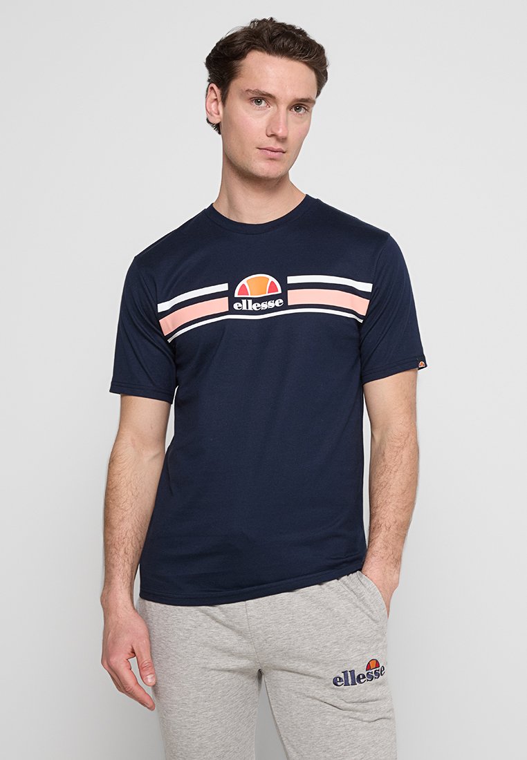 Mann trägt ein navyblaues ellesse T-Shirt mit rosa und weißen Streifen sowie hellgraue ellesse Jogginghose und steht mit einer Hand in der Tasche.