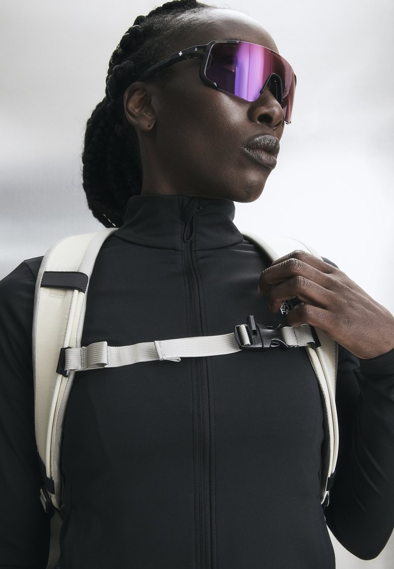 Persona con cabello trenzado que lleva gafas de sol deportivas reflectantes, chaqueta negra con cremallera y ajusta una correa blanca de mochila.