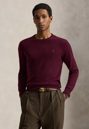 Junger Mann mit lockigem Haar, der einen burgunderfarbenen Pullover und braune karierte Hosen trägt, mit den Händen in den Taschen vor neutralem Hintergrund.