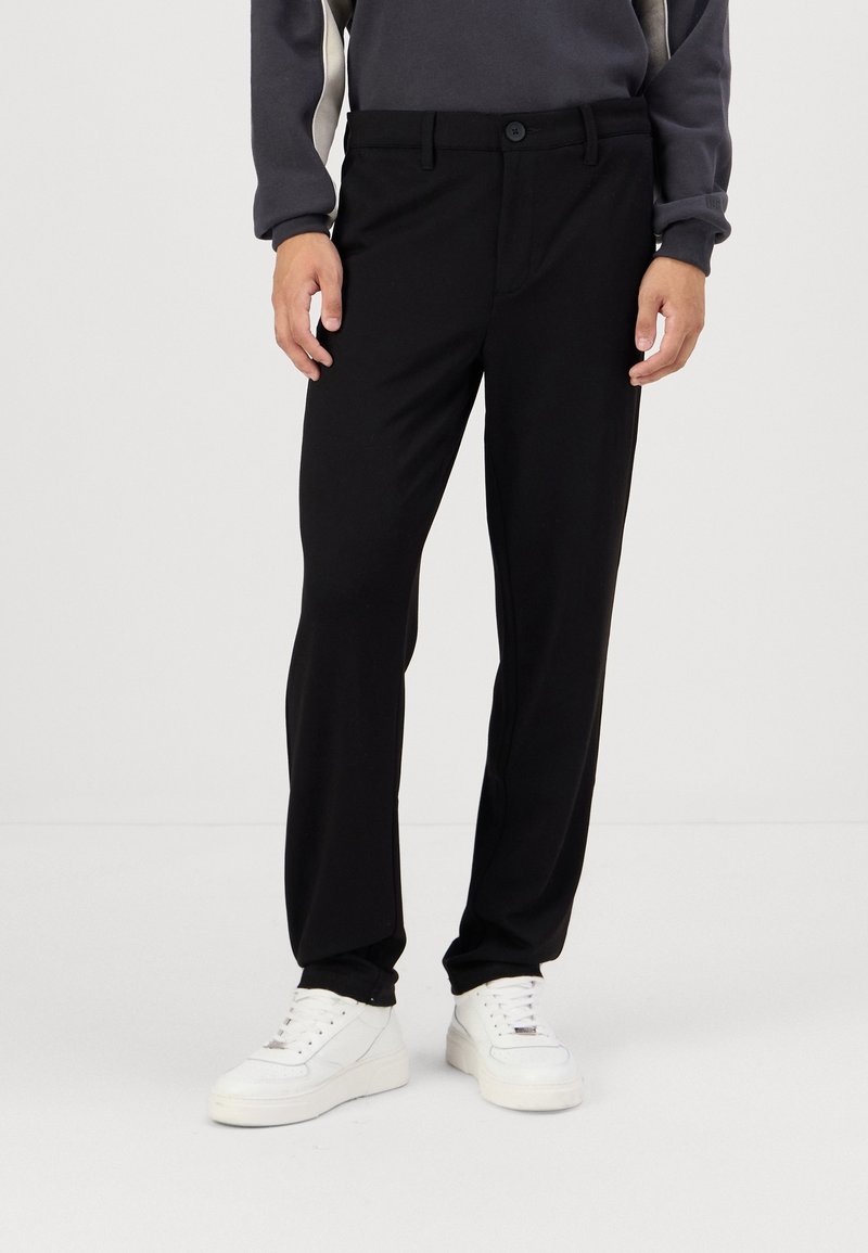 Homme portant un pantalon noir droit, des baskets blanches et un haut à manches longues gris, sur un fond blanc uni.