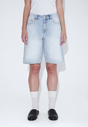 Persoon draagt lichtblauwe denim kniebroek, witte sportsokken, zwarte instapschoenen en een wit shirt tegen een effen witte achtergrond.