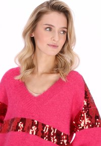 Roze gebreide trui met een V-hals, met horizontale strepen van rode pailletten op de mouwen. Zachte textuur en een comfortabele, losse pasvorm.