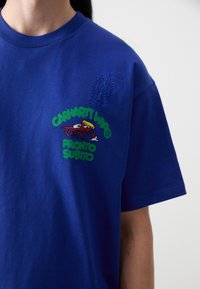 Kék pamut póló, zöld és piros grafikai designnal, rajta a "CARHARTT WIP" és "PRONTO SUBITO" feliratokkal, rövid ujjakkal és kerek nyakkivágással.