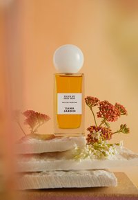Bouteille de parfum en verre clair avec un capuchon rond blanc, remplie de liquide ambré. L'étiquette indique "TIGER BY HER SIDE, SANA JARDIN." Accentuée de petites fleurs.