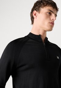 Crna zip-up majica s kratkim rukavima, s teksturnim naglascima na ramenima i malim logom na prsima. Glatka tkanina.
