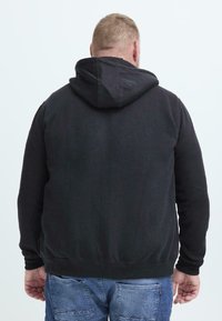 Uomo che indossa un hoodie nero e jeans blu, visto di spalle su uno sfondo chiaro.