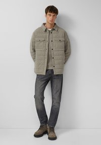 Beige gewatteerde jas met voorzakken over een licht sweater, gecombineerd met donkere jeans en bruine veterschoenen, staand tegen een neutrale achtergrond.