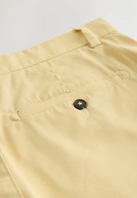 Détail du pantalon beige montrant une poche passepoilée arrière avec fermeture par bouton noir et une passants de ceinture au-dessus de la taille.
