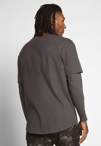 Pelēks garām piedurknēm T-krekls ar brīvu piegriezumu, apaļu kakla izgriezumu un mīkstu tekstūru; slāņota konstrukcija ar kontrastējošu īso piedurkni virs garās piedurknes.