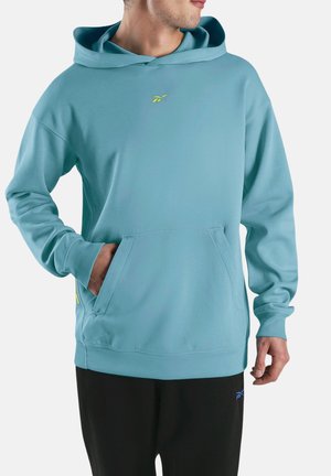 Mann trägt hellblauen Kapuzenpullover mit Kängurutasche und schwarze Hose, Hände teilweise in der Tasche, seitlich stehend.