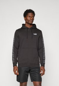Zwarte hoodie van katoen, met een kangoeroezak en een capuchon met trekkoord. De zijkanten van de mouwen hebben een bedrukt wit merklogo. Gecombineerd met zwarte shorts.