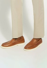 Brown leather slip-on shoes with a textured finish, white rubber sole, and decorative stitching along the edge. Worn with light beige trousers.