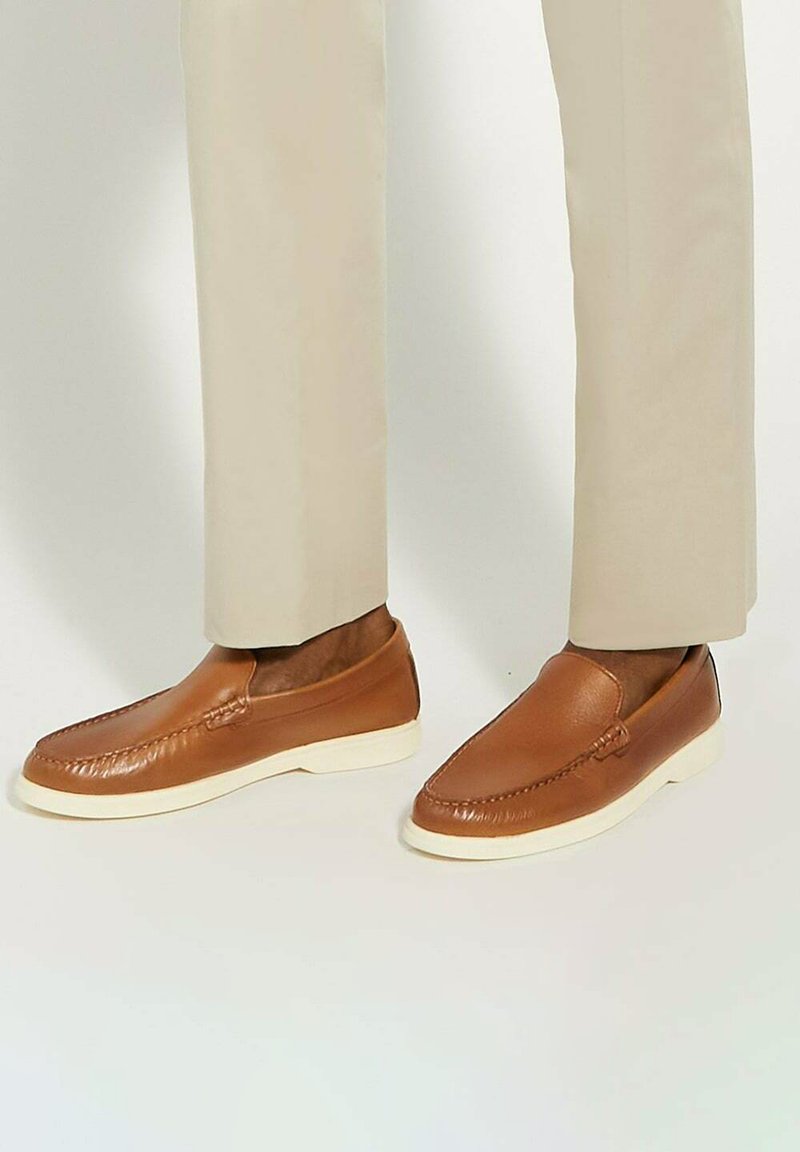 Brown leather slip-on shoes with a textured finish, white rubber sole, and decorative stitching along the edge. Worn with light beige trousers.