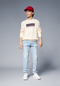 Felpa di colore panna con logo "Tommy HILFIGER", jeans azzurri, cappellino rosso e sneakers bianche. Design semplice con una vestibilità casual.