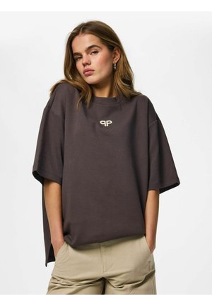 Jeune femme aux cheveux mi-longs portant un t-shirt oversize marron foncé avec un petit logo, pantalon beige, mains dans les poches, expression neutre.