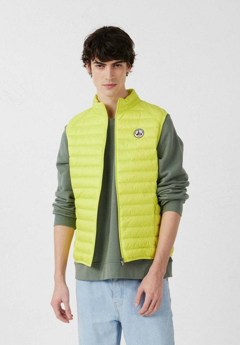 Limoengroene puffer-vest met ritssluiting en hoge kraag. Heeft een doorgestikt patroon en een logopatch op de borst. Gedragen over een grijze sweatshirt.