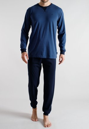 Mann in marineblauem Langarmoberteil und passender Hose, steht barfuß vor einem schlichten hellen Hintergrund.