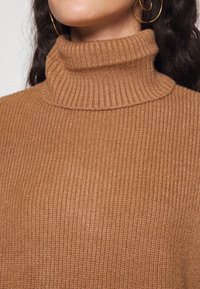 Brun ribbad polokragetröja med hög krage. Materialet är mjukt med en stickad yta och subtil stretch. Guldörhängen är synliga.