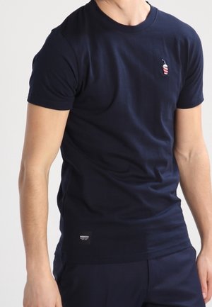 Uomo che indossa una maglietta aderente blu navy a manica corta con un piccolo ricamo a strisce rosse e bianche di una bevanda sul petto.