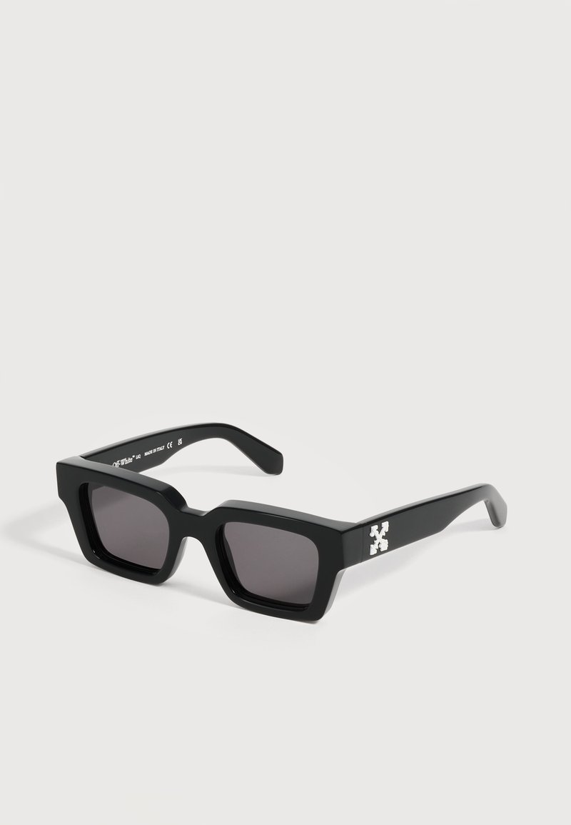 Lunettes de soleil rectangulaires noires avec des montures épaisses et des verres foncés, arborant un logo croix blanche sur les branches, posées sur une surface claire.