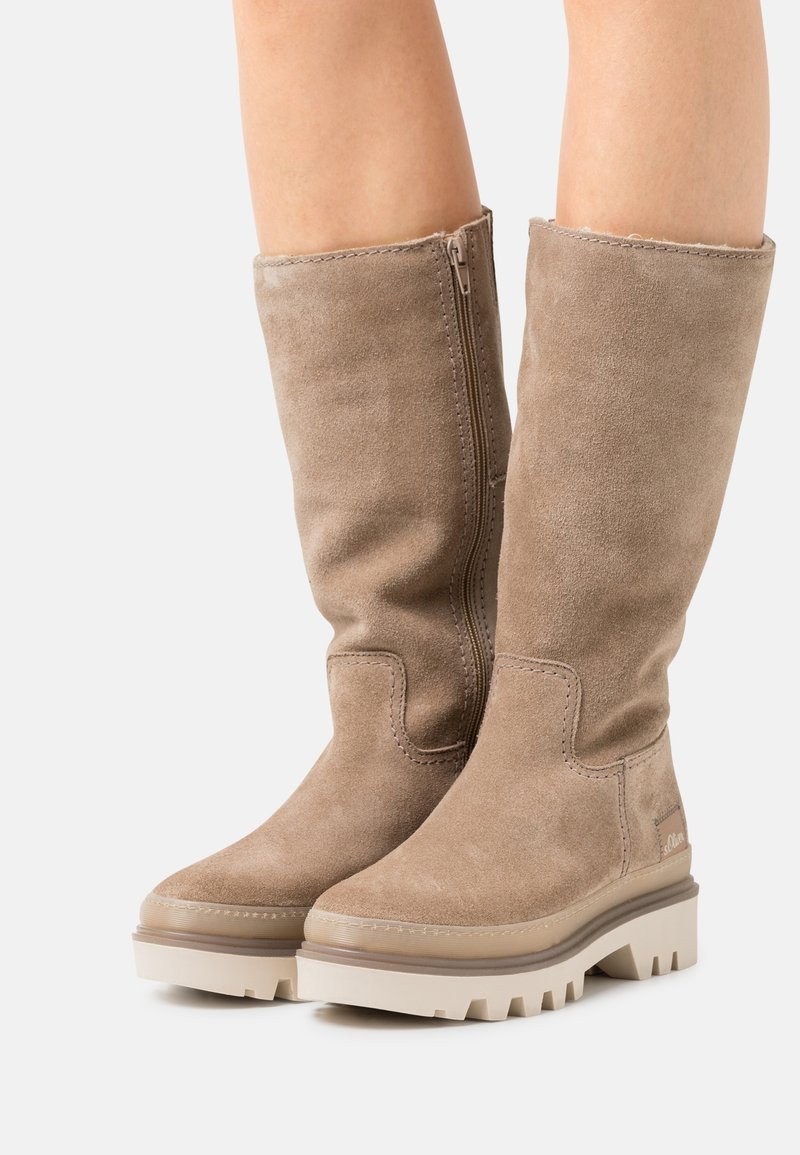 Bottes montantes en daim beige avec semelles épaisses à crampons et fermeture éclair latérale, portées sur des jambes nues, présentées sur un fond clair uni.