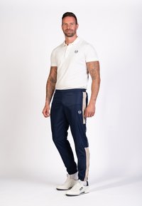 Marineblaue Jogginghosen mit beigen Seitenstreifen, elastischem Bund und einem Logo-Patch, kombiniert mit einem weißen Poloshirt und weißen Sneakern.