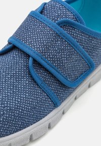 Niebieski tekstylny but z zapięciem na rzep, charakteryzujący się teksturowaną powierzchnią i wykonaną z miękkiego szarego materiału podeszwią. Miękka wewnętrzna wyściółka w kolorze niebieskim.