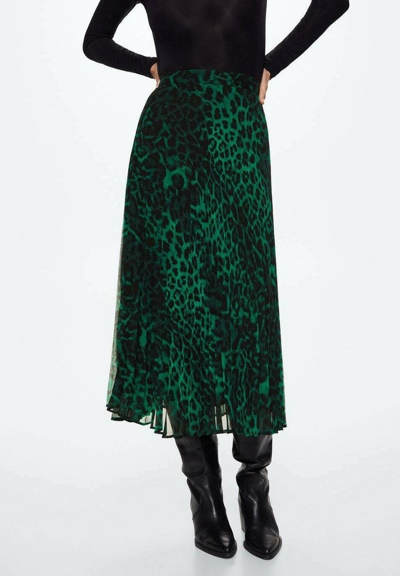 Femme portant une jupe midi imprimé léopard vert et noir, un haut noir à manches longues et des bottes noires à talons hauts pointues, debout les mains sur les hanches.
