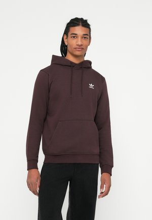 Ung mann med flettehår iført en mørk maroon Adidas hettegenser og svarte bukser, som står foran en enkel hvit bakgrunn.