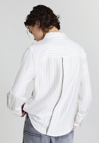 Camicia bianca a maniche lunghe con righe verticali, dotata di una zip sul retro in evidenza e tessuto testurizzato. Vestibilità comoda, orlo curvo.