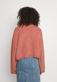 En person med lockigt hår som bär en loose fit, ribbad, korallrosa tröja och en högmidjad blå denimkjol, vänd bort mot en enkel bakgrund.