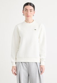 Felpa bianca con scollo rotondo, polsini e orlo a coste, caratterizzata da un piccolo logo verde ricamato sul petto. Indossata con pantaloni grigi.