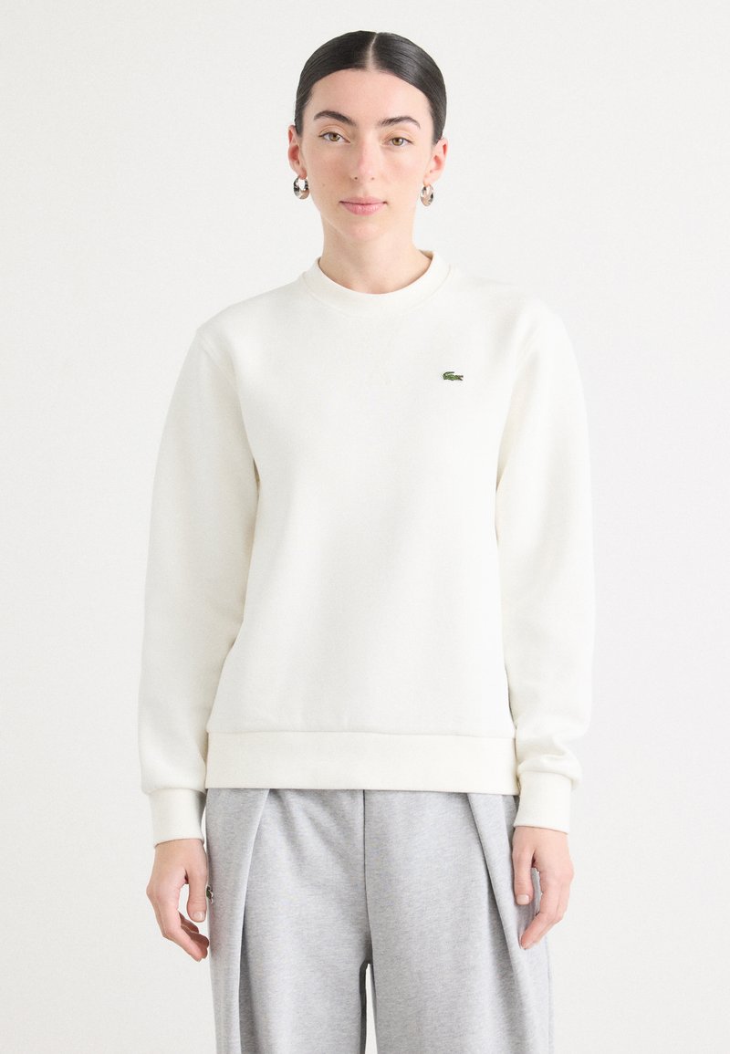 Felpa bianca con scollo rotondo, polsini e orlo a coste, caratterizzata da un piccolo logo verde ricamato sul petto. Indossata con pantaloni grigi.