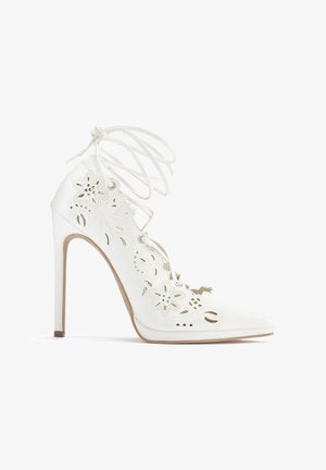 White high-heeled shoe with lace-up design, cutout floral pattern, pointed toe, and smooth texture. Stiletto heel adds height.