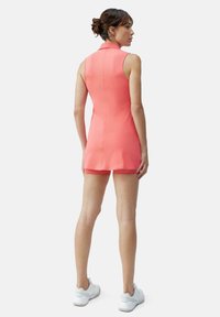 Korallkrāsas bez piedurknēm tērps ar pieguļošu dizainu un apkakli, kas raksturojas ar gludu tekstūru un aizmugurējo rāvējslēdzēju. Komplementēts ar baltām kedām.