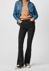 Jeans de mezclilla negros acampanados combinados con un suéter de cuello alto de canalé beige y una camisa de mezclilla azul. El atuendo incluye zapatillas negras con acentos blancos.
