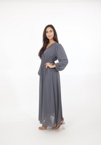 Mujer con cabello largo y oscuro, vestida con un vestido gris largo y sandalias transparentes de tacón alto, de pie con la mano en la cadera contra un fondo blanco.