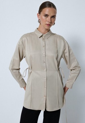 Vrouw met naar achteren gestyled haar draagt een beige blouse met lange mouwen en knopen, met zijkanten die gestrikt zijn, gecombineerd met een zwarte broek, staand tegen een effen achtergrond.