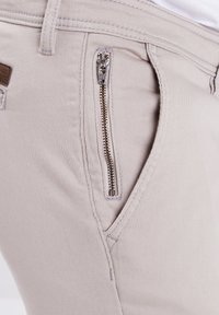 Beige katoenen broek met een zijzak met rits, voorzien van een zilveren rits. De stof heeft een soepele textuur en subtiele stikseldetails.