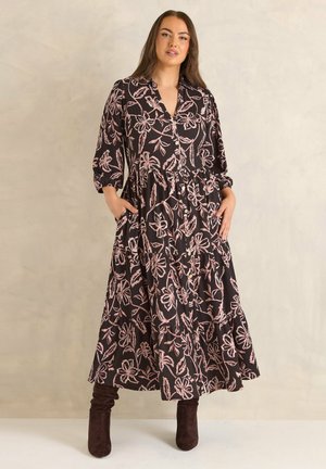 Femme portant une robe midi noire à motif floral rose, boutonnée à l'avant, manches trois-quarts, mains dans les poches, et bottes marron foncé.