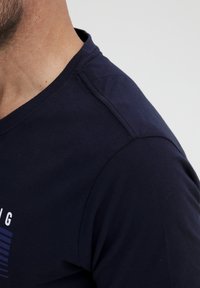 Tums zils kokvilnas T-krekls ar apaļu kakla izgriezumu, īsām piedurknēm un nelielu grafisko dizainu, kas attēlo horizontālas svītras uz priekšpuses.