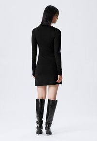 Juoda ilgomis rankovėmis suknelė su prigludusia silueto forma, lygi medžiaga, iki vidurio šlaunies ilgio, derinama su aukštais juodais batais. Rodomas nugaros vaizdas.