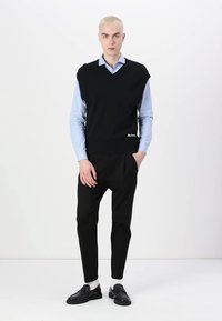 Svart uten ermeløs genser over en lys blå skjorte med krage, kombinert med svarte smale bukser og svarte loafers. Glatte teksturer, minimal design.