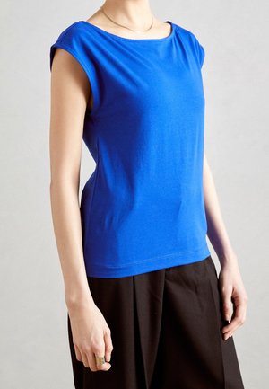 Woman wearing a bright blue sleeveless top with a round neckline paired with loose black pants against a neutral background.