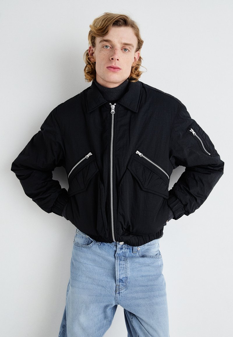Chaqueta bomber negra con bolsillos con cremallera, un cuello y puños elásticos. Lleva puesta sobre un cuello de tortuga y combinada con unos jeans de color azul claro.
